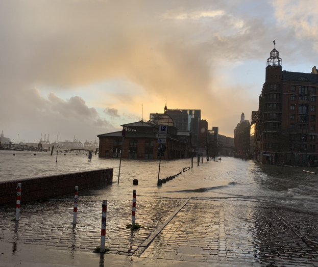 Eine überflutete Straße in Hamburg, Deutschland, mit Wasser, das die Straße, Strommasten, Schilder, Gebäude mit Fenstern, eine Brücke und einen bewölkten Himmel bedeckt.