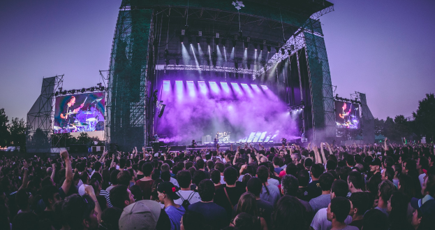 Eine große Menschenmenge versammelte sich auf einem Musikfestival, auf dem eine Bühne mit Musikern, die Instrumente spielen, im Mittelpunkt steht, beleuchtet von Lichtern und umgeben von Bildschirmen, mit einem Gebäude, Bäumen und einem klaren blauen Himmel im Hintergrund.