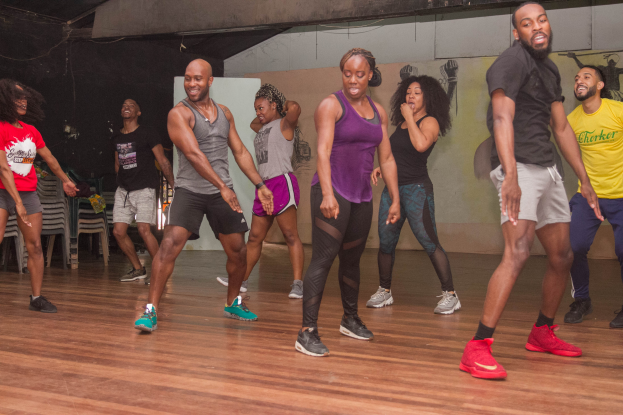 Eine Gruppe von Menschen in Turnschuhen tanzt in einem Studio mit einer bemalten Wand, Stühlen und anderen Gegenständen unter einer Decke mit Lampen und einem sichtbaren Boden.