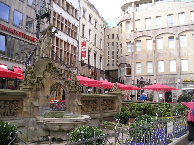 Eine Stadtansicht mit Gebäuden, Zelten, Pflanzen und Menschen im Hintergrund und einer Skulptur, einem Teich, einem Geländer und Pflanzen im Vordergrund.