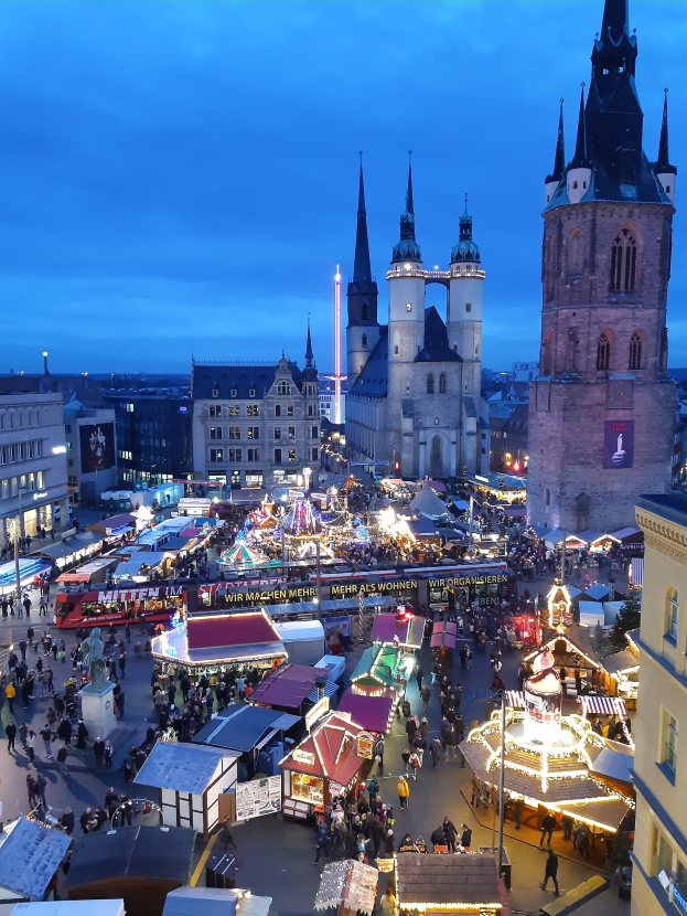 Ein nächtlicher Weihnachtsmarkt in Köln, Deutschland, mit Menschen, die auf dem Boden stehen und von den Lichtern der Stände und Gebäude beleuchtet werden, unter einem bewölkten Himmel.