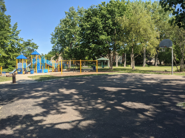 Ein Park mit einem Spielplatz in der Mitte, umgeben von Bäumen unter einem klaren blauen Himmel, mit einer Straße unten und einem Basketballkorb auf der rechten Seite.