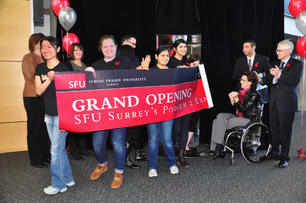 Eine Gruppe von Frauen hält ein Banner hoch und bewirbt ein Ereignis, mit zwei klatschenden Männern daneben und einer sitzenden, aufgeregten Frau im Rollstuhl vor einem schwarzen Vorhang und Ballons.
