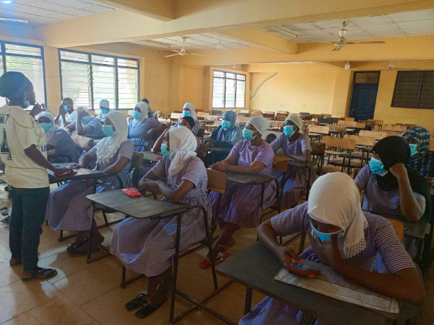 Ein Klassenzimmer in Nigeria mit Schülern, die Gesichtsmasken tragen und auf Bänken und dem Boden sitzen, mit Büchern und Gegenständen auf den Tischen, Fenstern, einer Tür, einer Wand, einer Decke mit Lampen und Ventilatoren im Hintergrund.