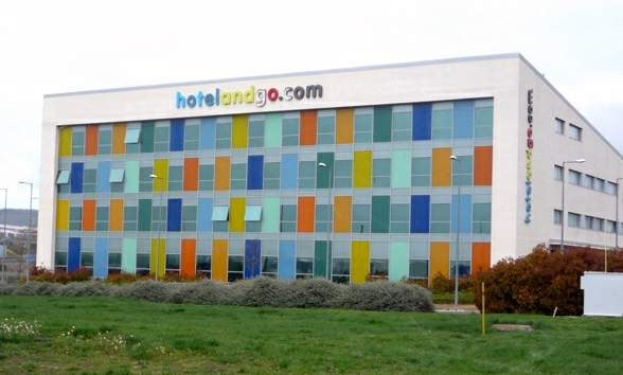 Großes modernes Hotelgebäude mit zahlreichen Fenstern, umgeben von Grünflächen, einem Zaun, Straßenlaternen, geparkten Fahrzeugen, fernen Hügeln und einem bewölkten Himmel, mit der Aufschrift "hotel and go" an seiner Fassade.