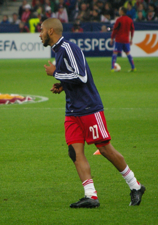 Ein Mann in einem blauen T-Shirt und roten Shorts läuft auf einem Fußballfeld, umgeben von sitzenden Zuschauern und einigen Werbetafeln, trägt Fußballschuhe.