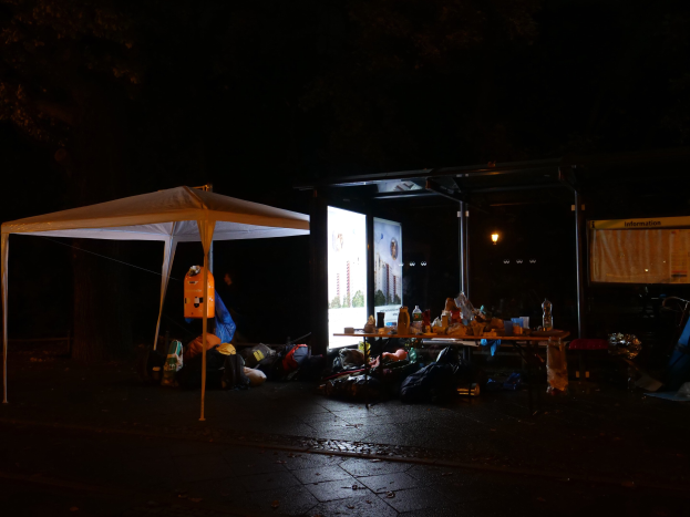 Ein Zelt auf einer Straße bei Nacht mit einem Tisch davor, auf dem Flaschen und andere Gegenstände stehen, umgeben von verstreuten Taschen, und ein beleuchteter Bildschirm im Hintergrund.