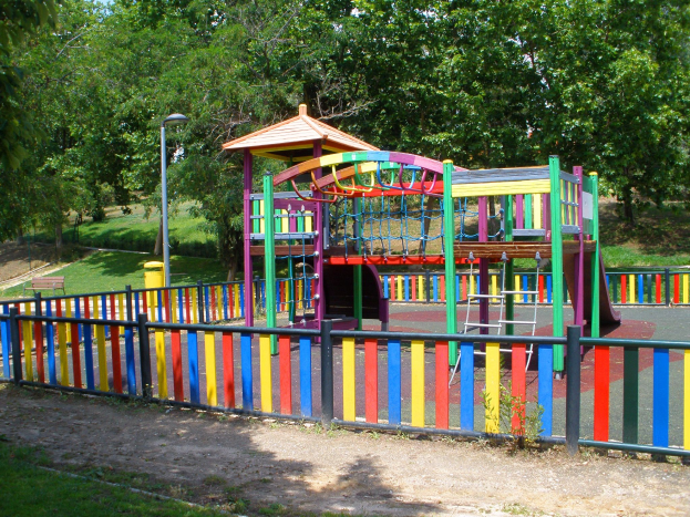 Ein Spielplatz mit einer bunten Spielstruktur in der Mitte, umgeben von einem Zaun, Pfosten, Laternen, Bänken, Gras, Pflanzen und Bäumen unter einem klaren blauen Himmel.