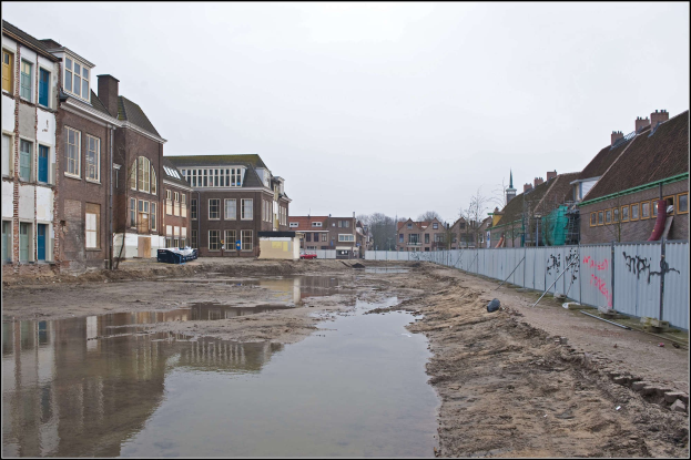 Flutstraßen in der Stadt mit Wasser auf dem Boden, ein Zaun auf der rechten Seite, Gebäude mit Fenstern auf der linken Seite, Bäume im Hintergrund und Himmel darüber.
