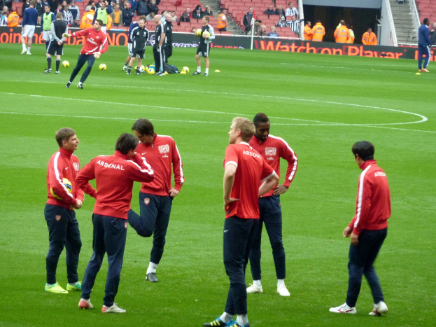 Eine Gruppe von Spielern auf einem grünen Feld mit Fußballen, während das Publikum auf Stühlen im Hintergrund sitzt.