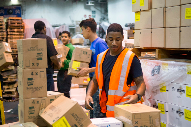 Männer in orangen Westen und Sicherheitswesten laden Kisten in ein Lager, mit Kartonagen auf der rechten Seite und einer Kette auf der linken Seite, unter Lampen an der Wand im Hintergrund.