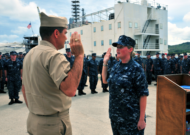 Ein Mann und eine Frau in Militäruniformen stehen nebeneinander vor einem Podium mit Flaschen und anderen Gegenständen, mit einem großen Schiff mit Fenstern, Geländern, Stufen und einer Fahne mit einem Mast dahinter und Hügeln und einem bewölkten Himmel im Hintergrund.