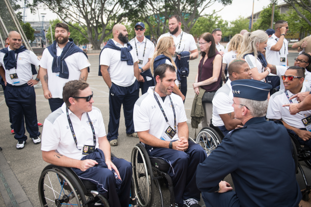 Eine Gruppe von Menschen im Rollstuhl, die vor einem Hintergrund aus Bäumen, Pfählen, Gebäuden und einem klaren blauen Himmel miteinander sprechen, wobei einige Mützen und Brillen tragen, was darauf hindeutet, dass sie bei den Paralympischen Spielen sind.
