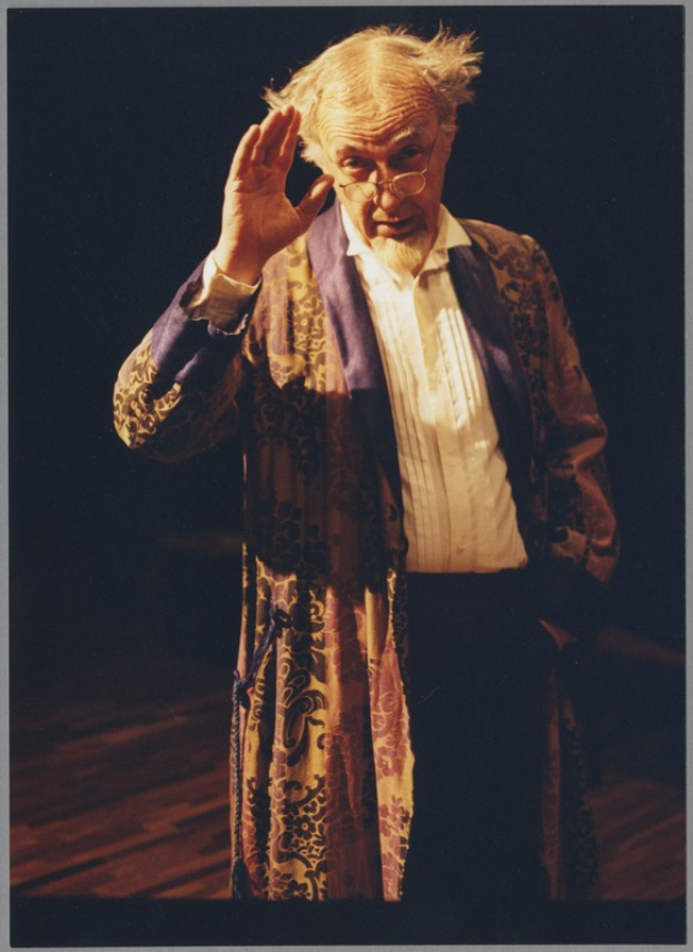 Ein Mann in einem Gewand und mit Brille steht auf einer Bühne mit dunklem Hintergrund und schaut ernst in die Menge.