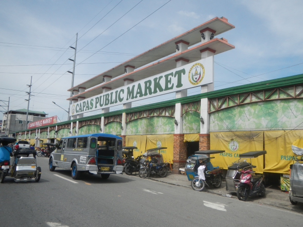 Lebendige Stadtstraße mit Autos, Motorrädern und Rikschas, die an dem Gebäude "Capas Public Market" vorbeifahren.