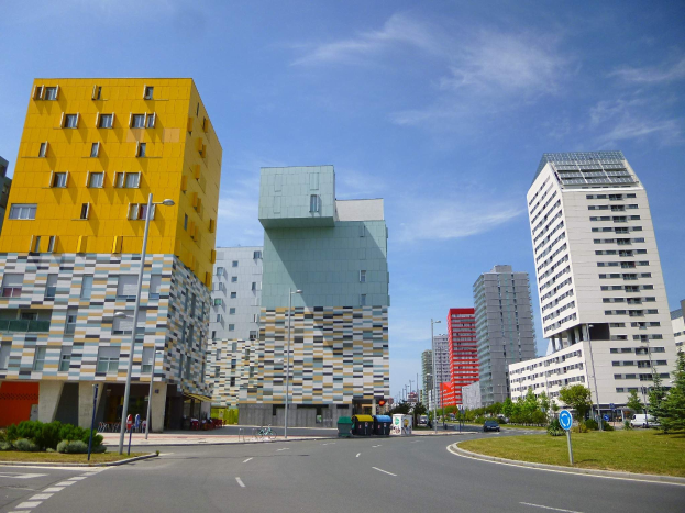 Eine Stadtstraße mit hohen Gebäuden der Universität Rotterdam, Straßenlaternen, Laternen, Fahrzeugen, Infotafeln, Grünflächen und einem bewölkten Himmel.