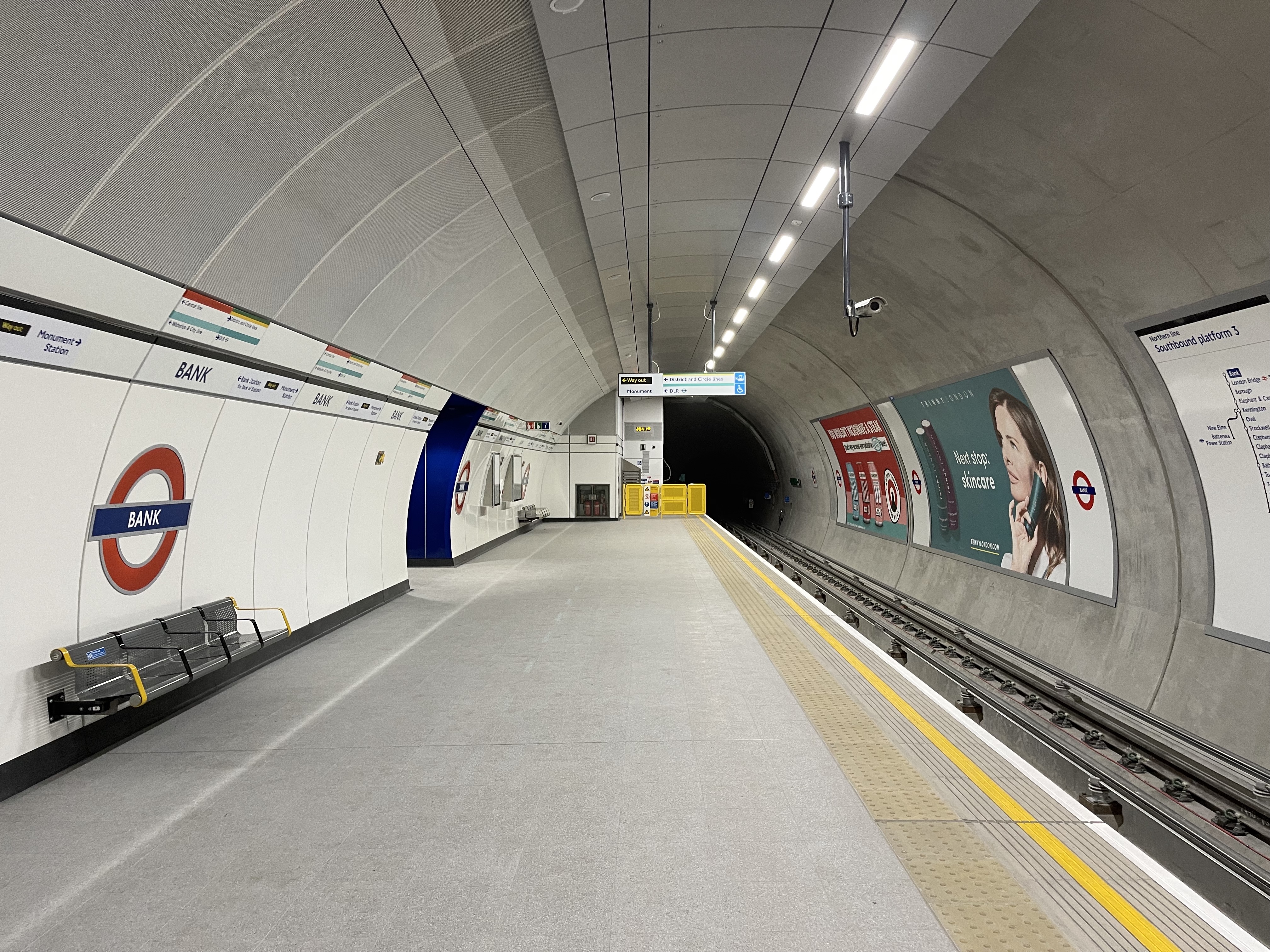 Eine U-Bahn-Station mit einem Zug auf den Gleisen, ein Bahnsteig mit Bänken, Tafeln mit Text und Bildern, Lampen an der Decke und eine Überwachungskamera.