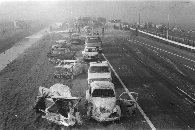 Ein Schwarz-Weiß-Foto von mehreren verunglückten Autos am Straßenrand bei Nacht, mit Straßenlaternen, einigen Menschen in der Nähe und Bäumen mit einem Gewässer im Hintergrund.