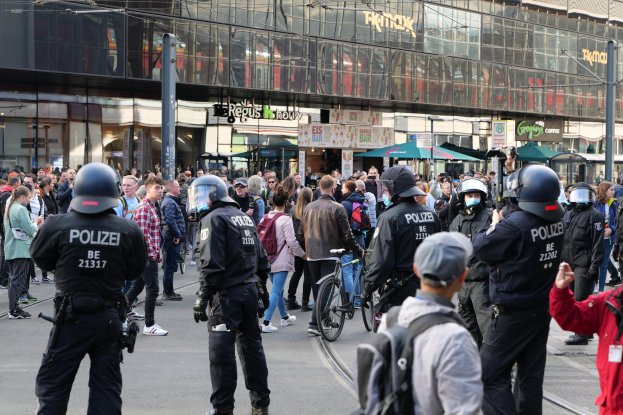 Eine Gruppe von Polizisten steht vor einer Menge auf der Straße, einige tragen Helme und haben Taschen oder Fahrráder dabei, im Hintergrund sind Gebäude, Laternenmäste und Schirme zu sehen.