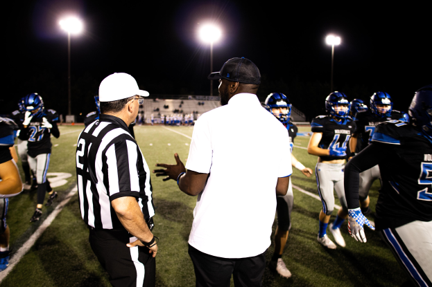 Ein Fußballtrainer in einer Mütze spricht mit seinem Helmträger-Team auf einem Feld bei Nacht, mit Leuchtmasten und dunklem Himmel im Hintergrund.