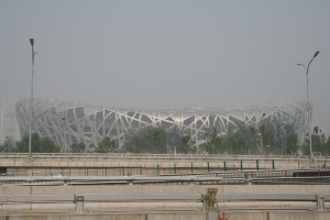 Olympiastadion in Peking mit Brücke, Geländern, Pfählen, Lichtern, Bäumen und einem Gebäude vor einem klaren blauen Himmel.