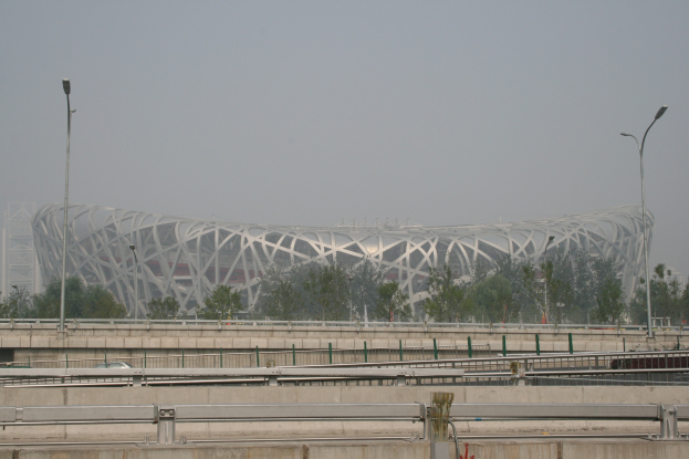 Olympiastadion in Peking mit Brücke, Geländern, Pfählen, Lichtern, Bäumen und einem Gebäude vor einem klaren blauen Himmel.