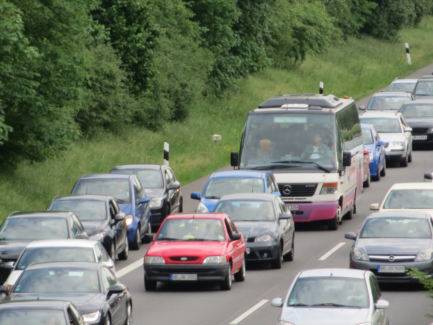 Verkehrsstau auf der Autobahn mit vielen Autos und einem Lieferwagen, Menschen in Fahrzeugen sichtbar, Bäume und Gras im Hintergrund.