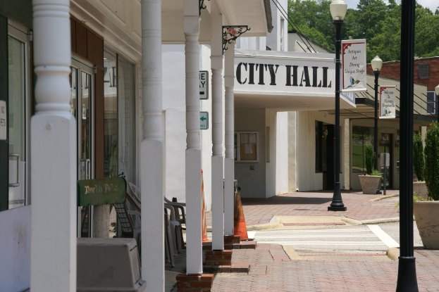 Ein Rathaus umgeben von Gebäuden, Straßenmasten, Straßenlaternen, Namensschildern, Kraftfahrzeugen, Absperrgittern, Stühlen, Topfpflanzen, Bäumen und einer bewölkten Himmel.