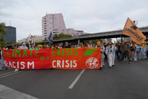 Eine Gruppe von Menschen marschiert eine baumbestandene Straße entlang und hält ein Banner mit der Aufschrift "Klimakrise ist eine Krise", mit Gebäuden und einer Brücke im Hintergrund unter einem bewölkten Himmel.