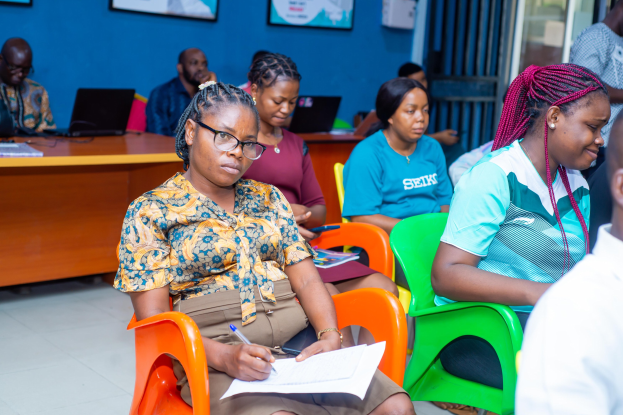 Eine Gruppe von Menschen, die auf Stühlen in einem Raum mit Laptops, Büchern und anderen Gegenständen auf Tischen sitzen, mit gerahmten Gegenständen an der Wand und einer Tür im Hintergrund.