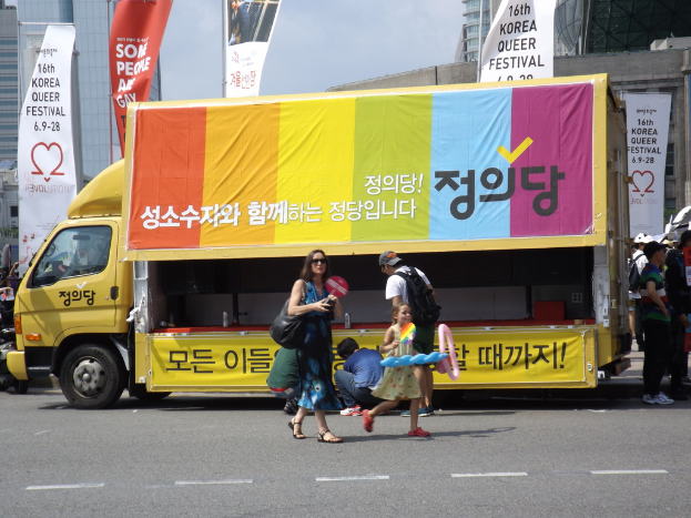 Eine belebte Stadtstraße mit Menschen, einem Motorrad und einem Lastwagen im Vordergrund, Schilder und Gebäude im Hintergrund unter einem sonnigen Himmel.