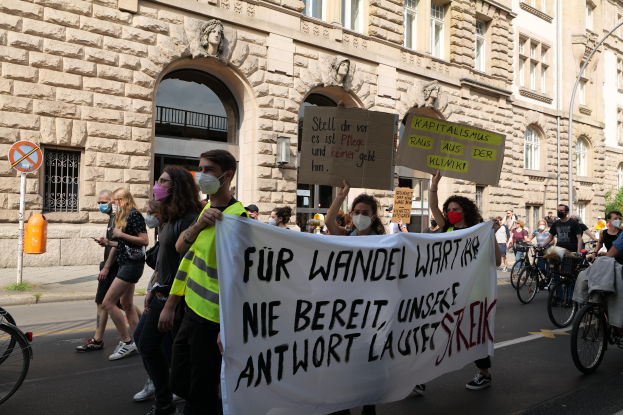 Eine Gruppe von Menschen marschiert auf einer Straße in Berlin, hält Schilder und Banner, während einige Fahrräder fahren, vor einem Gebäude mit Bögen, Säulen, Skulpturen und Bäumen.