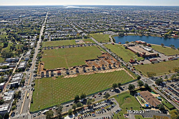 Luftaufnahme einer Stadt mit Gebäuden, Bäumen, Gras, Straßen mit Fahrzeugen, Wasser und Wolken, mit einem großen zentralen Feld, das für einen Park vorgesehen ist.