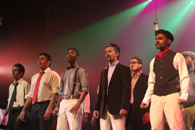 Eine Gruppe von M├Ąnnern auf einer B├╝hne stehend, die ein Drama auff├╝hren, mit gr├╝nen Scheinwerfern dahinter.