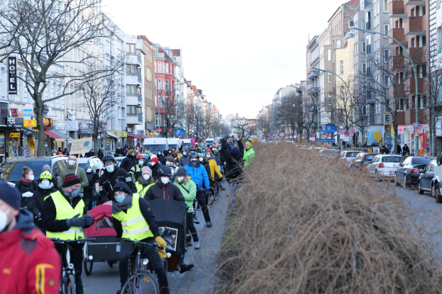 Eine große Gruppe von Menschen mit Masken und Sicherheitswesten auf Fahrrädern auf einer von Bäumen gesäumten Straße mit Gebäuden, Fahrzeugen und einem klaren blauen Himmel.