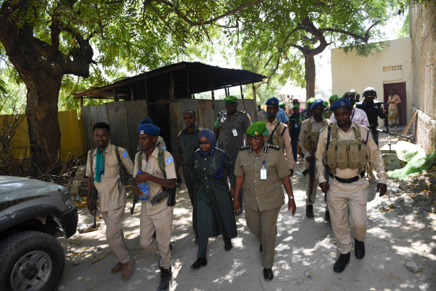 Gruppe von Polizisten in Uniform, die eine Straße entlanggehen, einige halten Waffen, mit Bäumen, Schuppen, Wänden und Gebäuden im Hintergrund.