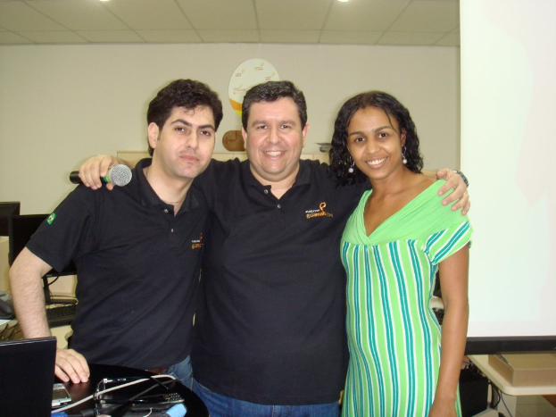 Zwei Männer und eine Frau, die lächeln und stehen, wobei einer der Männer ein Mikrofon hält, vor einem Tisch mit Mobilgeräten und einer Wand im Hintergrund.