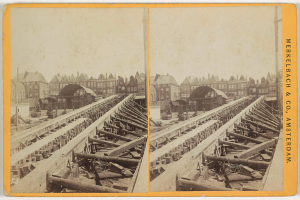 Ein altes Schwarz-Weiß-Foto einer Bahnschiene mit Gebäuden im Hintergrund, Text auf der rechten Seite lesend "Merkelbach & Co. Amsterdam" und verstreute hölzerne Gegenstände im Vordergrund.