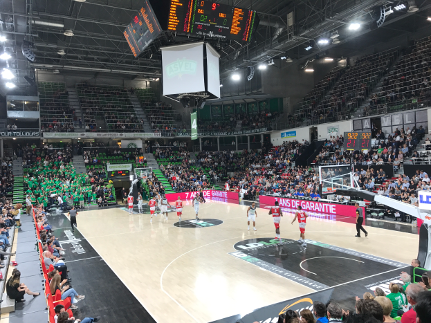 Ein Basketballspiel in einer großen Arena mit Zuschauern auf den Tribünen, Anzeigetafeln mit "Asvel vs Galatasaray" und einer Decke mit Lampen und einem Bildschirm.