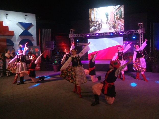 Eine Gruppe von Menschen, die auf einer Bühne vor einem großen Bildschirm tanzen, umgeben von einem sitzenden und stehenden Publikum, mit Blumentöpfen, Lichtern und anderen Gegenständen im Hintergrund.