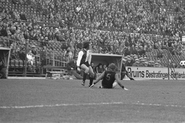 Ein Schwarz-Weiß-Foto eines Fußballspiels im Stadion, mit Spielern auf dem Feld und Zuschauern auf den Bänken im Hintergrund, mit sichtbaren Pfosten und Brettern.