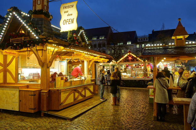 Ein nächtlicher Weihnachtsmarkt auf einem Kopfsteinpflasterstraßen mit Menschen, die beleuchtete Stände durchstöbern, umgeben von geschmückten Gebäuden, Bäumen und einem klaren Himmel.