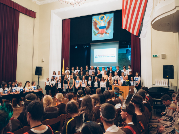 Eine Gruppe von Menschen steht an einem Podium mit einem Mikrofon während eines 2. Wochenende von Kunst und Kultur, flankiert von Fahnen und Lautsprechern, gegenüber einem sitzenden Publikum mit einem Bildschirm, der Text und ein Bild anzeigt, im Hintergrund.