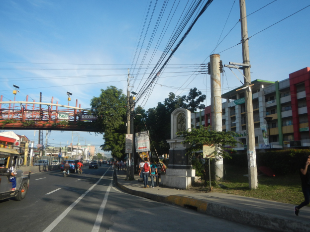 Eine belebte Stadtstraße mit Autos, Fußgängern, Gebäuden, einer Brücke und Bäumen unter einem bewölkten Himmel.