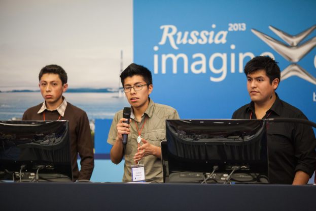 Drei Jungen stehen neben einem Schreibtisch mit zwei Computersystemen, ein Junge spricht in ein Mikrofon, alle tragen ID-Karten mit roten Markierungen, vor einer Wand mit einem Werbeplakat, auf dem "Russia Imagine 2013" steht.