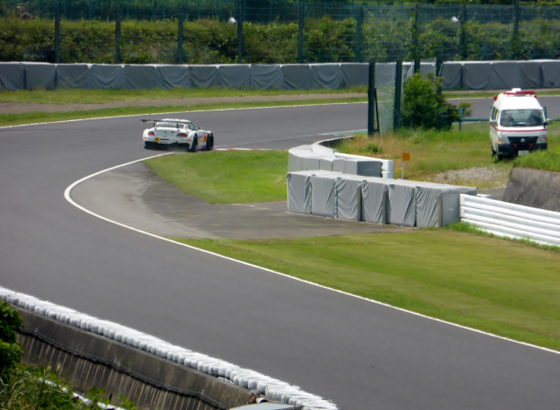 Ein Rennwagen fährt eine Rennstrecke entlang mit einem Polizeiauto im Hintergrund, umgeben von Grünflächen und Absperrungen, mit Text unten.