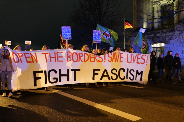 Eine Gruppe von Menschen marschiert eine von Bäumen gesäumte Straße entlang und hält ein Transparent mit der Aufschrift "Öffnet die Grenzen, rettet Leben, kämpft gegen Faschismus", mit einem Gebäude im Hintergrund.