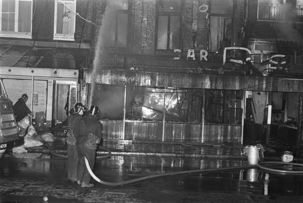 Feuerwehrleute in Schwarz-Weiß-Szene bei der Löschung eines Straßenrandfeuers, mit Menschen mit Rohren, einem geparkten Fahrzeug, verstreuten Gegenständen, einem Gebäude mit Fenstern, einem Schild mit Text und Drähten unter einem bewölkten Himmel.