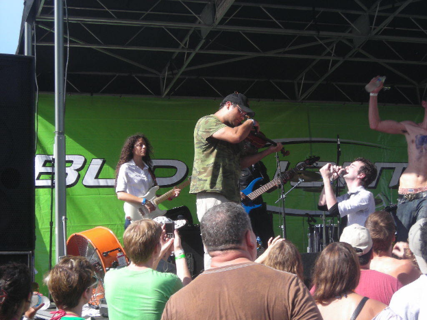 Eine Gruppe von Musikern, die verschiedene Instrumente auf einer Bühne spielen, während das Publikum vor einem Podium mit einer grünen Fahne im Hintergrund steht.