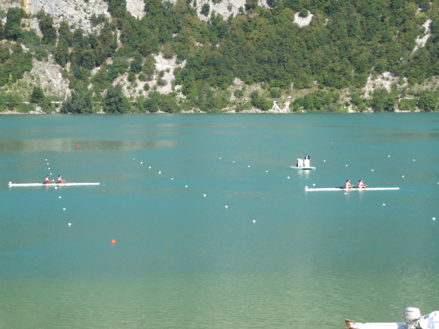 Eine Gruppe von Menschen, die auf einem Gewässer in einem Boot rudern, mit einem bewaldeten Hügel im Hintergrund und einem Boot am unteren Ende des Bildes, das auf einen Wettbewerb hinweist.